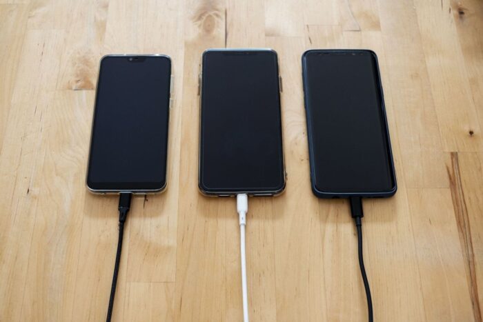 Tre smartphones med svarta skärmar laddas med kablar på ett träbord